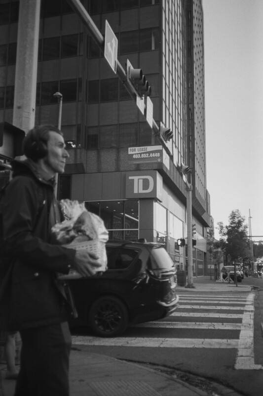 A person wearing headphones walks across a city street carrying bags. A black SUV waits at the crosswalk. A tall building with a "For Lease" sign is visible in the background. The image is in black and white.