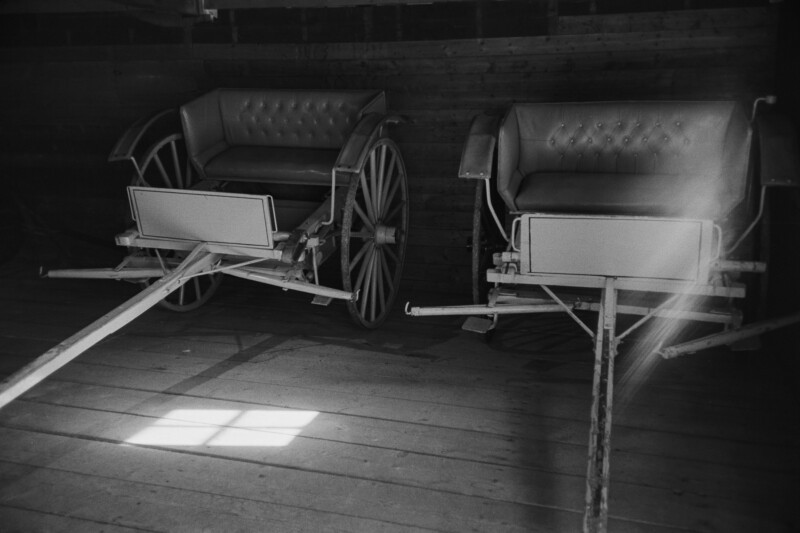 Black and white photo of two vintage wooden carriages with tufted seats inside a dimly lit room. Sunlight streams in, casting a bright rectangle on the wooden floor.