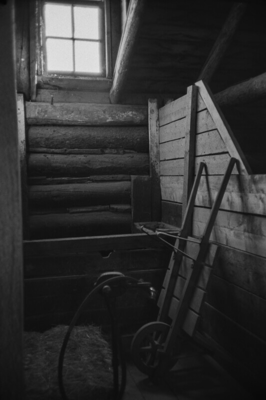 A black and white photo of an old, rustic wooden barn interior. A wheelbarrow and hose are leaning against a wooden wall. Light filters in through a small window, illuminating hay on the ground. Logs are visible in the wall construction.