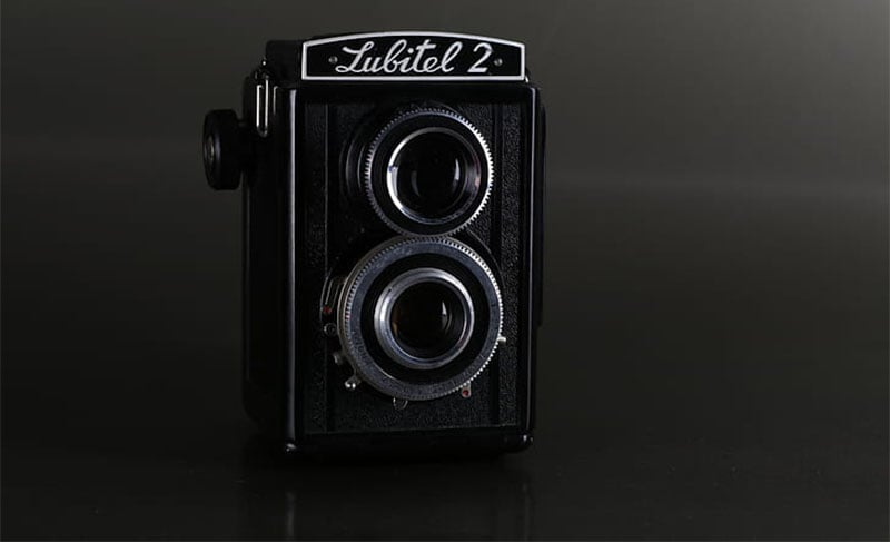 A vintage Lubitel 2 twin-lens reflex camera is set against a dark background. The black camera features two prominent lenses stacked vertically, with a textured body and the name "Lubitel 2" displayed on top.