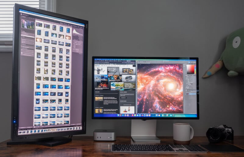 A dual monitor setup on a desk displays photo editing software. The left screen is vertically oriented, showing thumbnail images. The right screen features a galaxy image. A small computer, keyboard, mug, and camera are also on the desk.