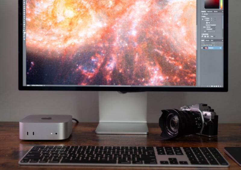 A computer monitor displays a vibrant, colorful galaxy image. On the desk are a compact Mac mini, a keyboard, and a vintage-style camera.