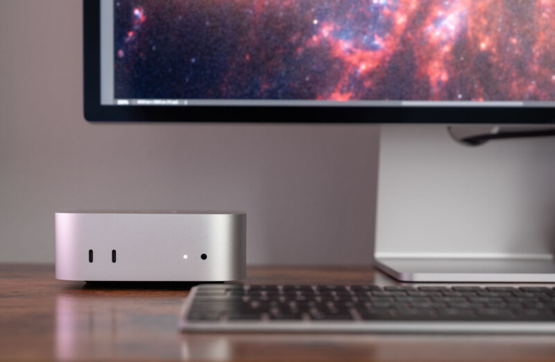 A sleek silver mini desktop computer sits on a wooden desk next to a monitor displaying a colorful galaxy image. A black keyboard is in the foreground. The setup appears modern and minimalist.