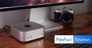 A sleek silver computer device with an apple logo is placed on a wooden desk in front of a large monitor. A digital camera and a keyboard are nearby. The text "PetaPixel Reviews" is overlaid on the image.