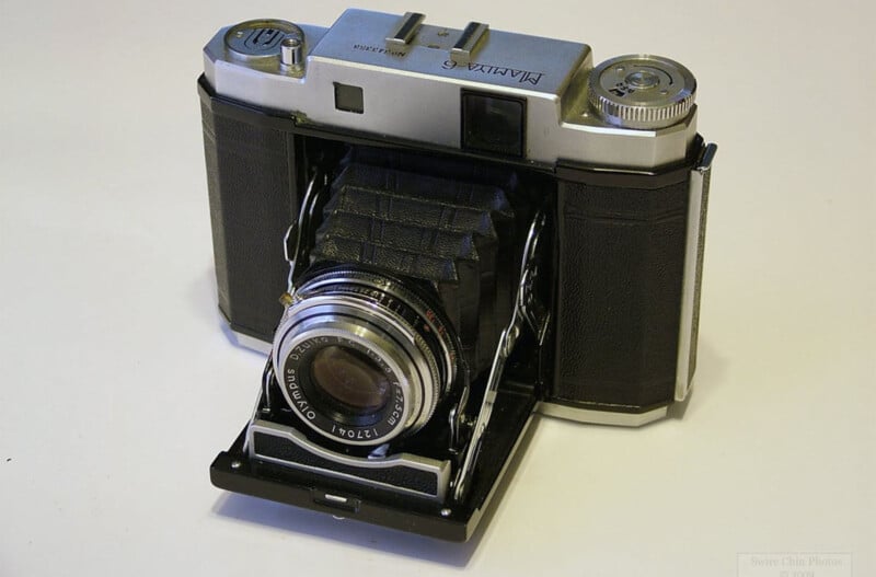 An old-fashioned Agfa camera with a folding bellows design, featuring a metallic body and black textured panels. The lens is prominently displayed as part of the classic, vintage setup.