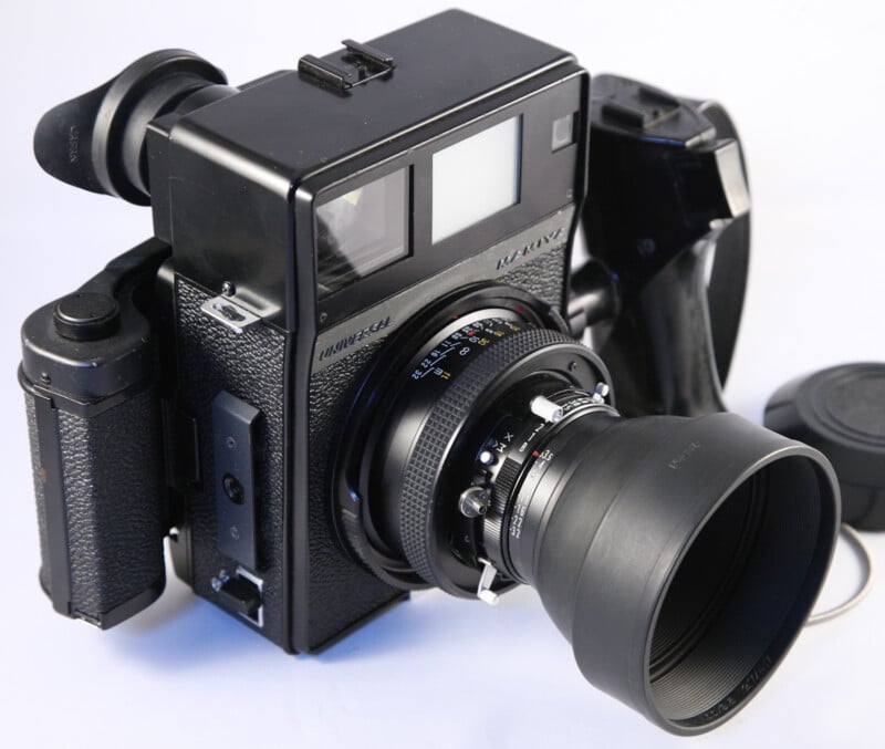 A vintage medium format camera with a large lens and viewfinder. The camera is black with textured grips and has multiple dials and a detachable lens cap nearby. It is set against a plain white background.