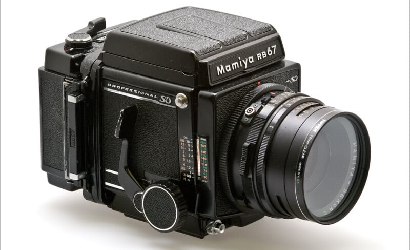 A black Mamiya RB67 medium format film camera with a large lens, displayed on a white background. The camera features robust design elements typical of professional photography equipment.