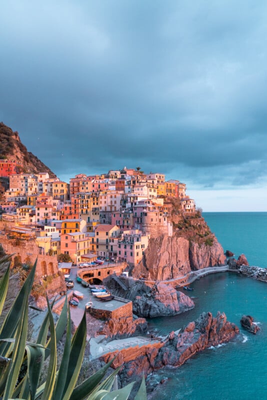 A picturesque seaside village with colorful buildings perched on a rugged coastline. The turquoise sea surrounds the rocky cliffs, while dramatic clouds fill the sky, capturing a serene and vibrant coastal scene.