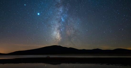 A breathtaking view of the night sky with the Milky Way galaxy prominently visible. The scene includes a silhouetted mountain range and its reflection in a calm body of water below. The sky is filled with numerous bright stars against a dark backdrop.