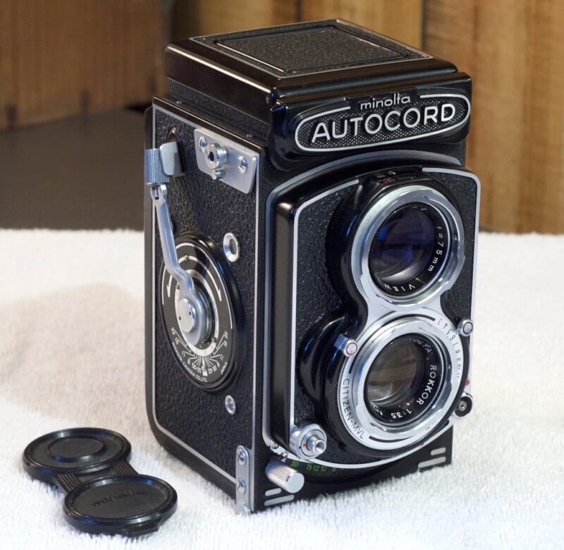 A vintage Minolta Autocord twin-lens reflex camera is displayed on a white surface. The camera has a retro design with metal and textured black surfaces. Two lens caps are placed nearby.