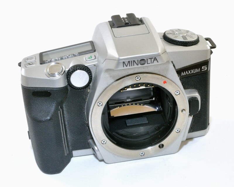 A silver Minolta Maxxum 5 film camera without a lens, showing the mirror inside the body. It has a black grip and various control dials and buttons on the top, with the brand and model name visible.