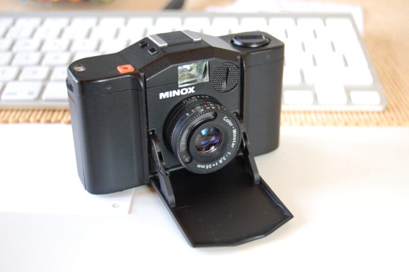 A black Minox film camera with a textured lens cap is resting open on a white surface. In the background, a blurred keyboard is partially visible.