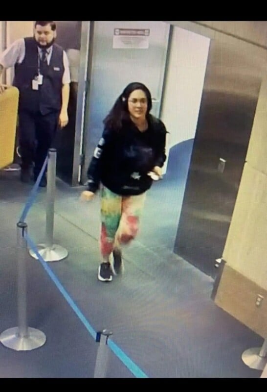 A woman wearing a black jacket and colorful rainbow pants walks quickly past a cordoned-off area in a building. A man in a uniform follows behind her.