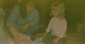 A woman and a young girl sit on the floor. The woman is holding a colorful book, showing it to the girl. The room is dimly lit, and there's a stack of colorful blocks in the background.