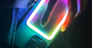 A hand holding a smartphone with a glowing rectangular light on its back. The light emits a spectrum of neon colors, including green, blue, and purple, adding a vibrant and futuristic effect to the scene.