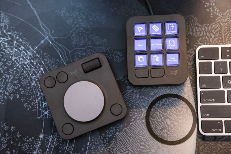 A photo of two compact Logitech input devices on a desk. One device has a large circular dial and a small joystick, while the other features several illuminated buttons with various icons. Part of a keyboard is also visible on the right side of the image.