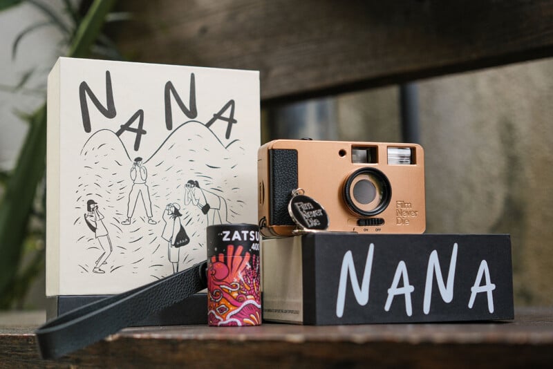 A vintage-style camera with "Film Never Die" text sits on a wooden surface. Next to it is a box displaying "NANA" and an illustrated mountain scene. A colorful film canister labeled "ZATSU" is also present.