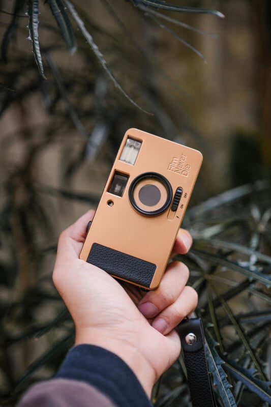 A person holding a vintage-style beige camera with a leather grip. It is surrounded by green, leafy plants. The camera is turned to show the lens and viewfinder.