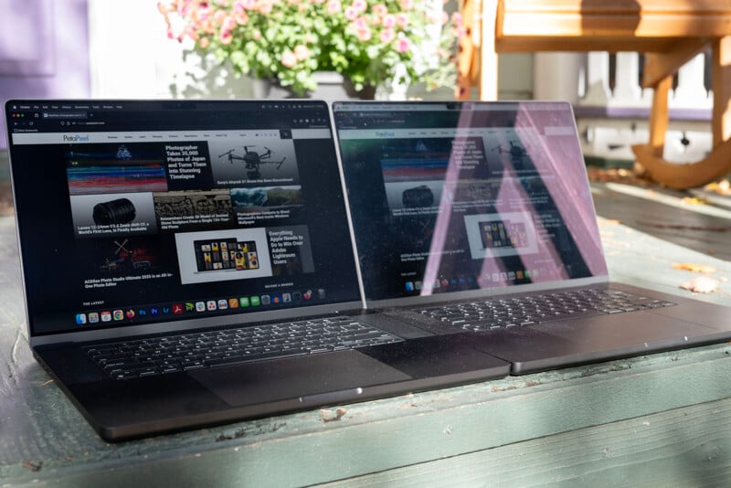 Two open laptops displaying the same webpage are placed on a green wooden table outdoors. The screens reflect light, and pink flowers can be seen in the background.