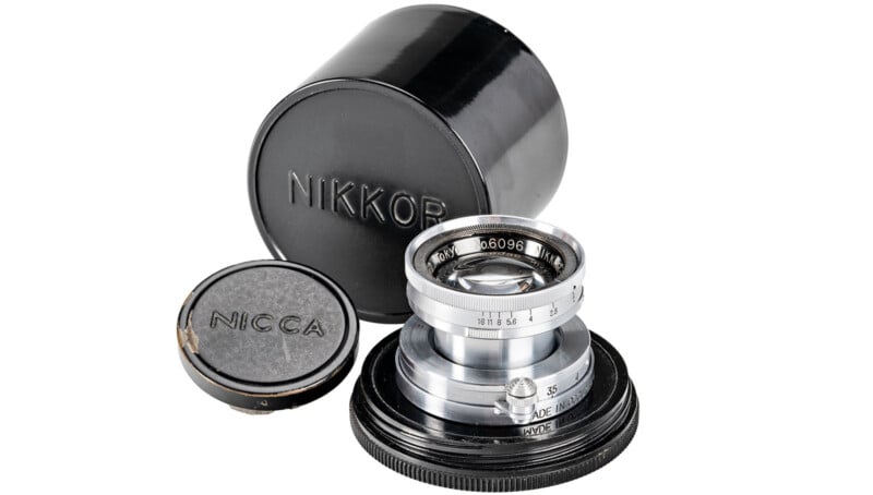 A vintage Nikkor camera lens with a silver finish is displayed standing upright. It is accompanied by a black lens cap marked "Nicca" and a cylindrical black lens case. The lens is set against a white background.