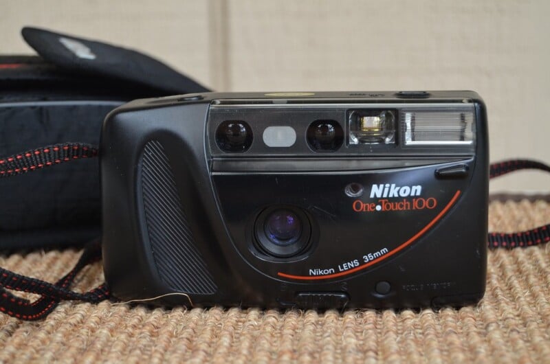 A Nikon One Touch 100 camera with a 35mm lens sits on a textured surface. The camera is black with red accents, showing a simple design with a built-in flash. A black case with a strap is partially visible in the background.