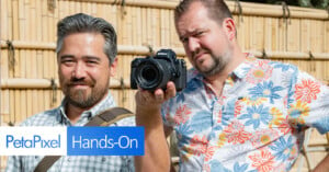 Two men are outdoors, both smiling. One holds a camera pointed towards the viewer. They stand in front of a bamboo fence, with a logo in the corner that reads "PetaPixel Hands-On.