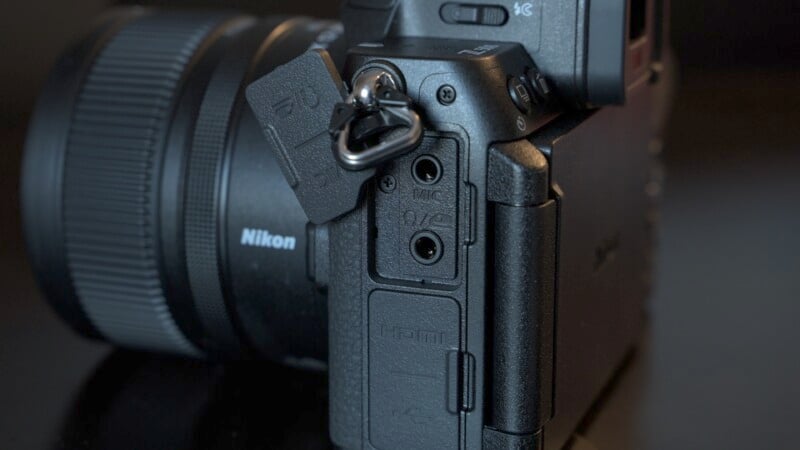 Close-up view of a Nikon camera showing the side ports. The lens is visible on the left, and the HDMI and microphone ports are uncovered, with a strap attached to the camera body.