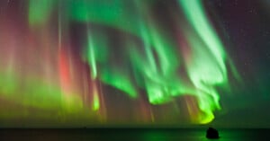 A breathtaking display of the Northern Lights illuminates the night sky with vibrant green and red hues. The auroras ripple elegantly over a darkened landscape, reflecting softly on a calm body of water below.