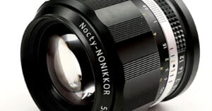 A camera lens labeled "Nocty-NONIKKOR 5" with a large reflective front element. The lens features a focus ring with ridged texture for grip and an aperture ring marked with f-stops from f/1.2 to f/16. The lens is shown on a plain white background.