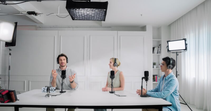 Three people sit at a table in a bright studio, wearing headphones and speaking into microphones. They are engaged in a conversation, surrounded by professional lighting and audio equipment.