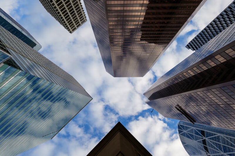 Upward view of modern skyscrapers with reflective glass facades converging towards a cloudy blue sky, creating a geometric pattern. The scene captures a sense of height and urban perspective.