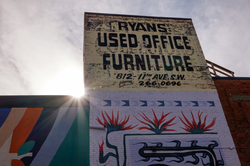 A sunlit mural on the side of a building showing a large vintage-style sign for "Ryans Used Office Furniture" with the address and phone number. Below the sign, there's colorful abstract artwork with floral elements.