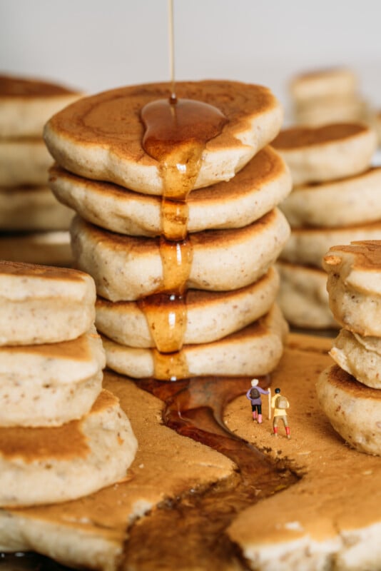 A stack of pancakes with syrup pouring down, surrounded by more pancakes. Two tiny human figures stand on a pancake, appearing to walk towards the syrup waterfall.