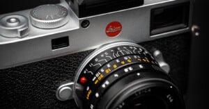 Close-up of a Leica camera focusing on the lens with detailed markings and the distinctive red Leica logo on the camera body. The textured black grip and metallic finish are visible, highlighting the precision and elegance of the design.