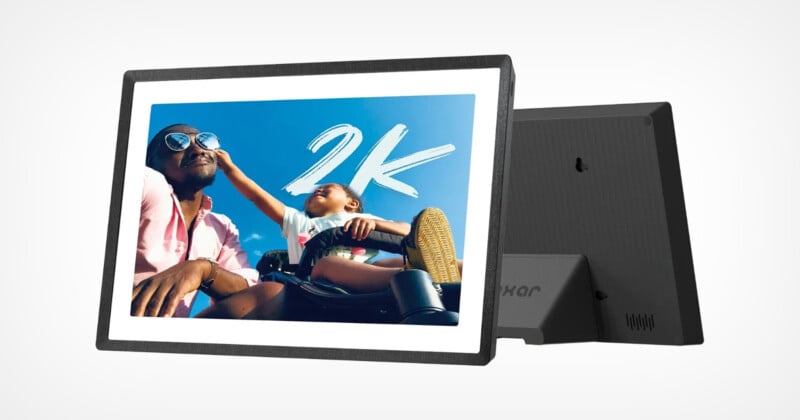 A digital photo frame displaying an image of a person and a child smiling against a clear blue sky. The child extends an arm playfully toward the person. "2K" is written in bold white letters on the image. The frame has a simple, modern design.