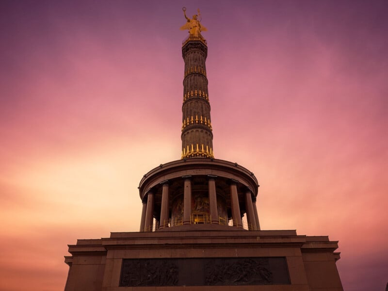 The image shows the Berlin Victory Column at sunset, with a vibrant pink and purple sky. The column is illuminated, highlighting its ornate details and the gilded statue of Victoria on top.