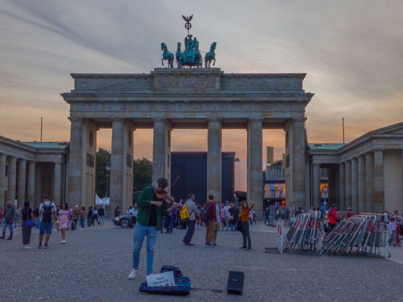 A street musician plays a violin in front of the Brandenburg Gate at dusk. People in the background are walking and taking photos. The sky has a mix of soft pastel colors as the sun sets.