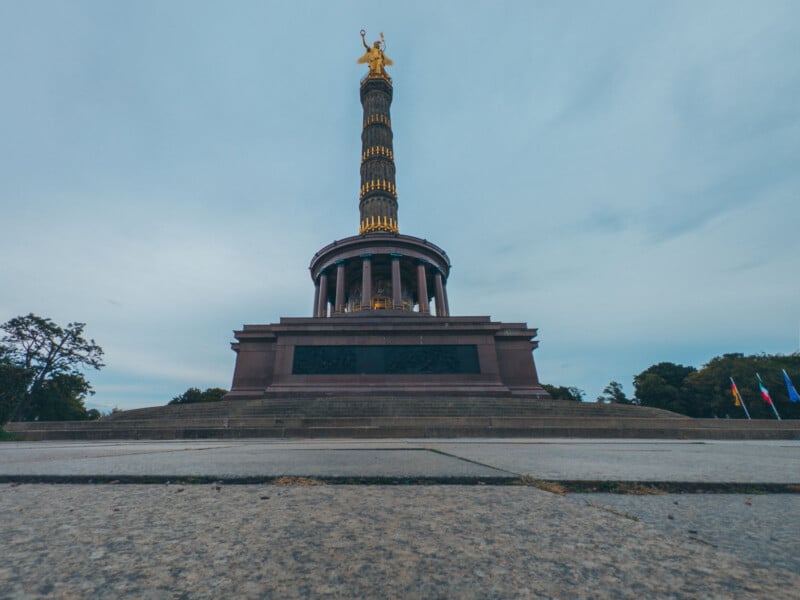 Low-angle view of the Victory Column in Berlin, set against a cloudy sky. The towering column is adorned with a golden statue on top. Trees and flags are visible at the base, adding color to the scene.