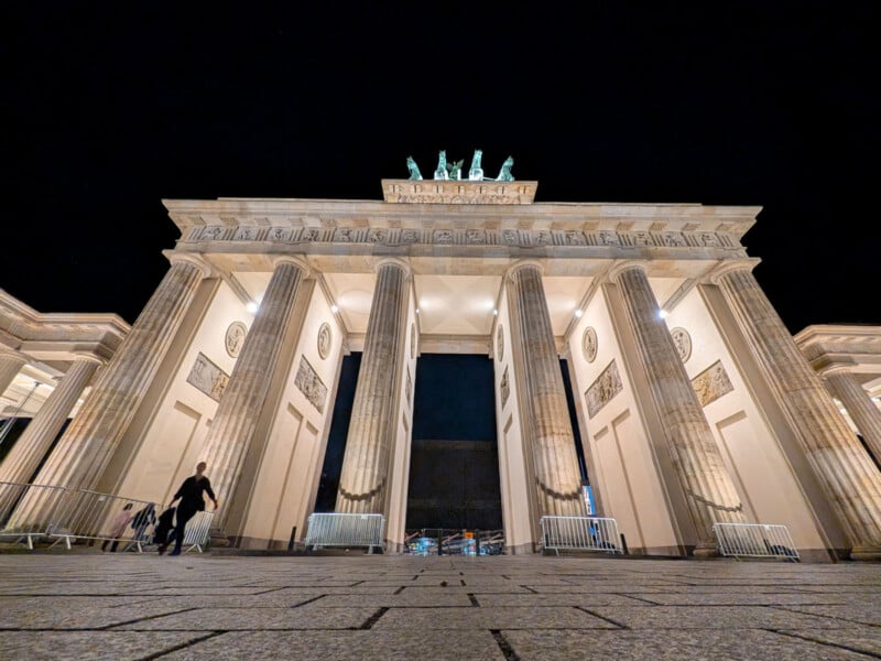 The image shows an upward view of the illuminated Brandenburg Gate in Berlin at night. The iconic neoclassical monument is adorned with a chariot statue on top, and a person is walking nearby on the left side.