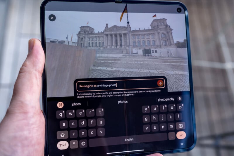 A person holds a foldable smartphone displaying a photo-editing app. The app shows an image of a large, historic building and a text box with the words "Reimagine as a vintage photo." A virtual keyboard is visible below.