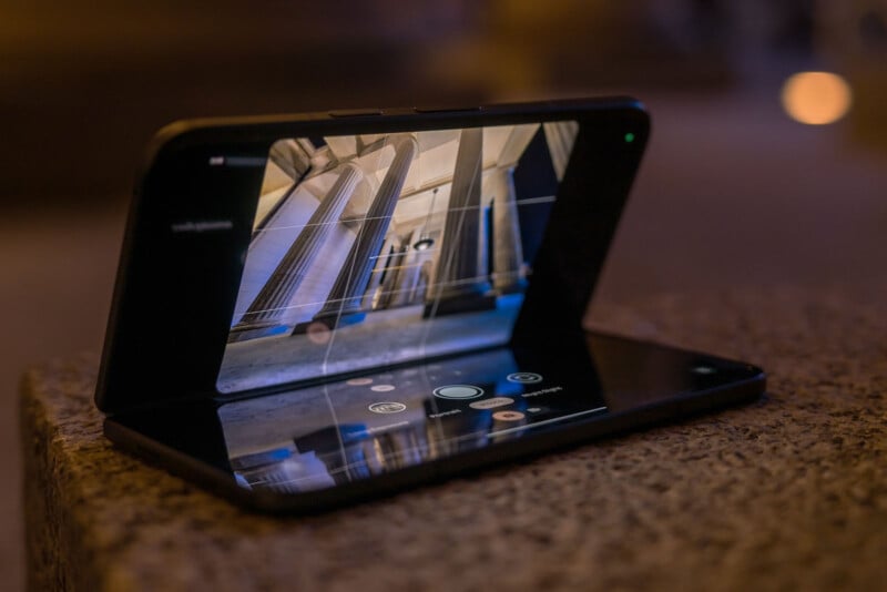 A foldable smartphone in flex mode displays a photo of columns on its screen, positioned on a textured stone surface. The dimly lit background includes blurred lights, creating a night-time ambiance.