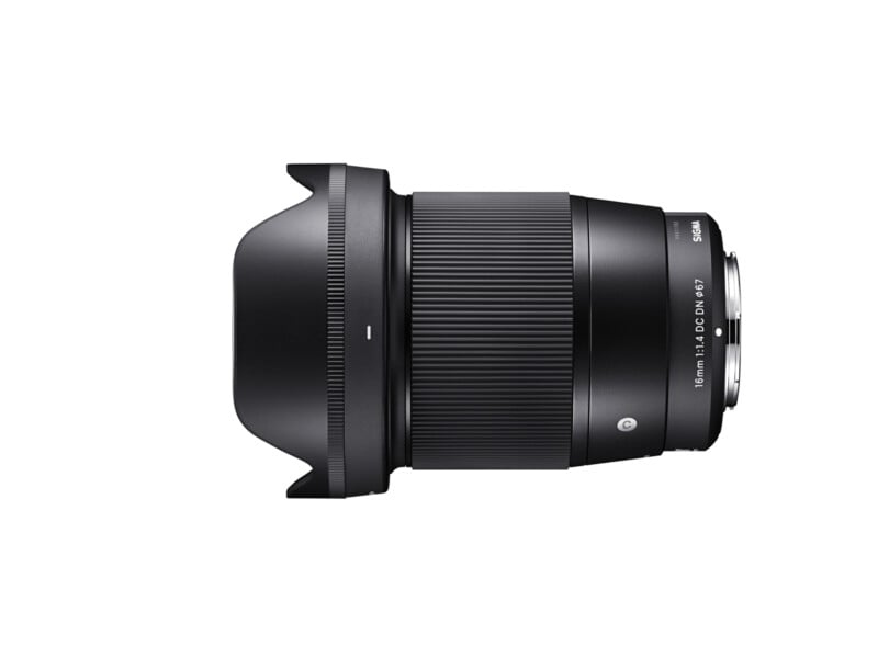 A black camera lens with a wide aperture, viewed from the side, showcasing a metal mount base and ribbed focus ring. The lens includes a lens hood, and markings on the lens barrel read "16mm 1:1.4 DC DN 67 Sigma.