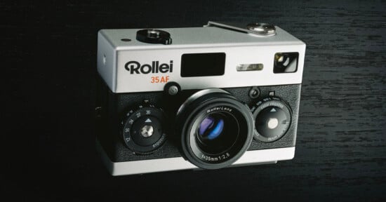 A vintage Rollei 35AF film camera sits on a dark wood surface. The camera features a silver body with black textured grips, a prominent lens, and various dials for settings. The brand name "Rollei" is visible in bold letters on the front.