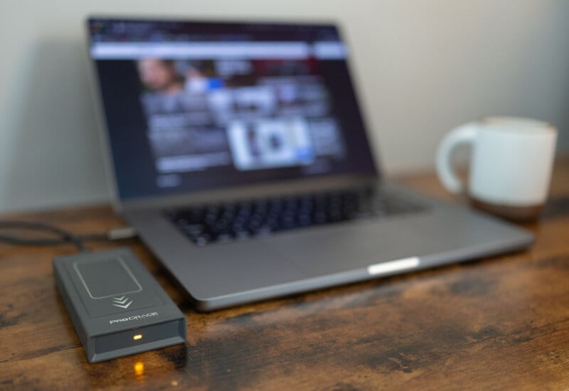 A laptop sits on a wooden desk with its screen displaying a blurred webpage. Next to the laptop is a Pinnacle brand device with an illuminated indicator. A white mug is placed near the laptop, all set against a plain background.