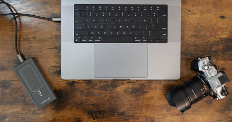 A laptop with a closed screen and a visible keyboard is placed on a wooden surface. An external hard drive is connected to the laptop via a cable, and a digital camera rests nearby, partially showing its lens.