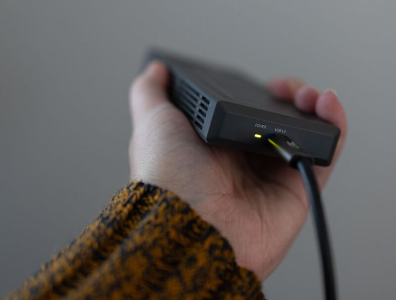 A person holds a small, rectangular external hard drive in their hand. The drive has a light indicator showing that it is powered on. A USB cable is connected to the device. The person is wearing a brown and black patterned long-sleeve top.