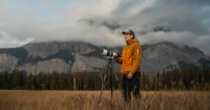 A person in an orange jacket stands in a grassy meadow with a camera on a tripod. In the background, misty mountains rise against a cloudy sky, partially shrouded in fog.