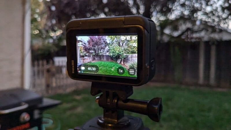 A camera on a tripod is focused on a backyard, capturing a view of green grass and trees through its display screen, which shows recording settings like 4K/30 fps. The background shows a wooden fence and a slightly blurred garden scene.