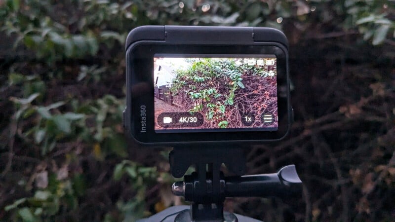 A camera screen displaying a viewfinder image of a pathway bordered by green foliage and a hedge. The screen shows settings like 4K/30fps. The camera is mounted on a tripod against a backdrop of dense greenery.