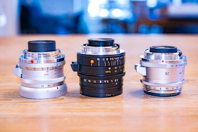 Three camera lenses of different sizes and colors are positioned on a wooden table. The lenses have various markings and settings visible, with two silver lenses on the sides and one black lens in the center. The background is softly blurred.